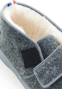 Grijze wollen slipper met een zachte binnenvoering, klittenbandsluiting en een geprofileerde buitenkant met een vlakke zool. Aangevuld met kleurrijke labels.