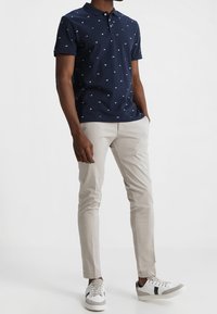 Polo bleu marine avec motifs géométriques blancs, associé à un pantalon slim beige clair et des baskets blanches avec des accents gris.