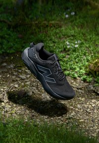 Schwarzer Sportschuh mit einem atmungsaktiven Mesh-Obermaterial, einer strukturierten Sohle, geometrischen Akzenten und einer abgerundeten Zehenpartie, vor einem natürlichen Hintergrund.