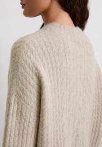 Beżowy sweter dzianinowy z teksturalnym wzorem w warkocze, wykonany z miękkiej wełny, o luźnym kroju, z okrągłym dekoltem i opadłymi ramionami.