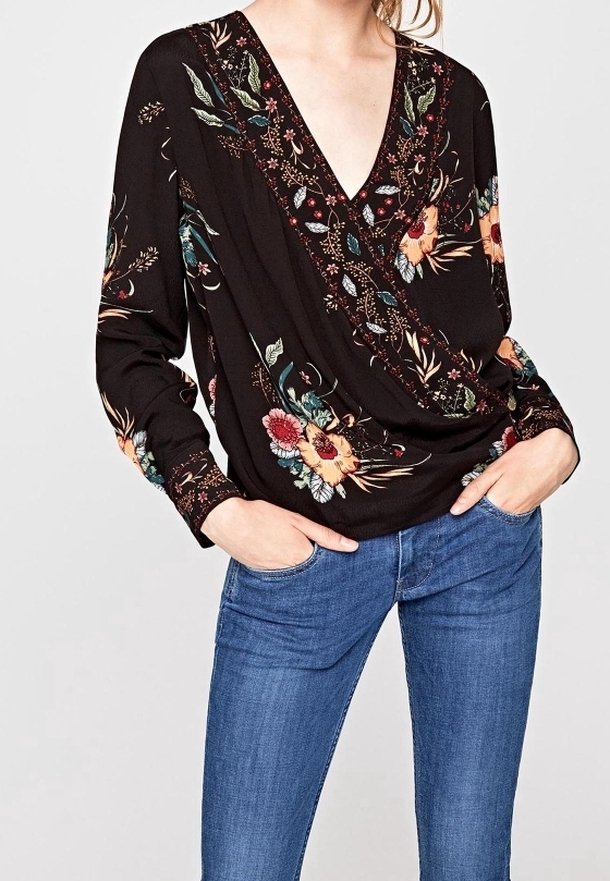 Blouse noire à manches longues avec un décolleté en V profond, une broderie florale aux couleurs vives et une coupe ample et drapée. Associée à un jean bleu.