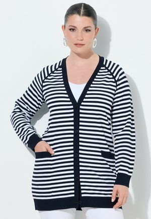 Vrouw met donker haar in een staart, draagt een zwart-wit horizontaal gestreepte cardigan met zwarte biezen, knopen aan de voorkant en voorzakken.