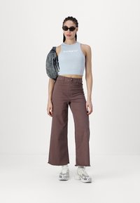 Top court bleu clair avec texte blanc, pantalon large marron et baskets argentées. Un bras porte un sac en denim. Lunettes de soleil rondes noires portées.