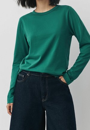 Vrouw draagt een nauwsluitend groen shirt met lange mouwen, ingestopt in een broek met hoge taille, donker en wijde pijpen, met haar hand in de zak.