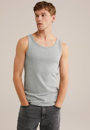 Junger Mann mit hellbraunem Haar, der ein graues ärmelloses Shirt und dunkelgraue Jeans trägt, die Hände in den Taschen, schaut zur Seite.