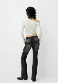 Top bianco a maniche lunghe con taglio off-shoulder abbinato a jeans svasati grigio scuro. L'outfit presenta una texture liscia e dettagli minimi.