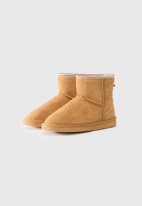 Bottes de cheville en daim beige avec un bout arrondi, une tige courte et un bord supérieur décoré de coutures. Semelle en caoutchouc visible en bas.