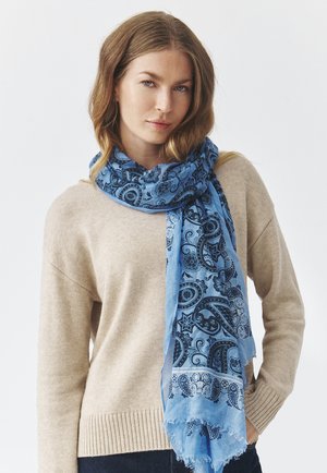 Écharpe bleu clair avec des motifs paisley noirs, bords à franges, en tissu doux et léger, drapée autour du cou sur un pull beige.