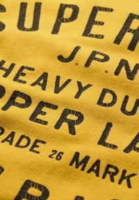 Żółta bawełniana koszulka z dużym, zniszczonym czarnym napisem: "SUPER J.P.N. HEAVY DUTY SUPER LAB GRADE 26 MARK." Miękka w dotyku, casualowy design.