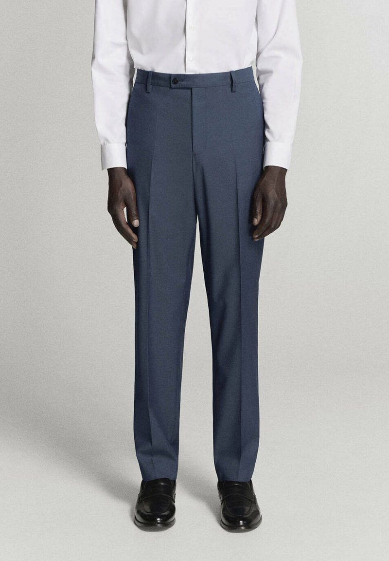Homme portant un pantalon formel bleu marine, des chaussures noires habillées et une chemise blanche à manches longues boutonnée, debout devant un fond uni.