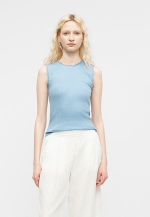 Mladá žena s dlhými blond vlasmi, oblečená v bezrukávovom svetlomodrom pletenom topu a voľných bielych nohaviciach, stojaca pred jednoduchým svetlým pozadím.