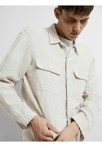 Lichtbeige linnen shirt met een kraag, twee borstzakken, knoopsluiting en lange mouwen. De stof heeft een subtiele textuurafwerking.