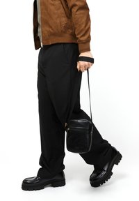 Borsa a tracolla in pelle nera con superficie testurizzata, chiusura con zip superiore e tracolla regolabile, tenuta da una persona in giacca marrone e pantaloni neri.