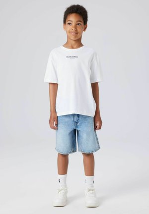 Poiss seisab, kandes valget "Studio edition" t-särki, helesiniseid teksapükse, valgeid sokke ja valgeid tossusid ühevärvilise tausta ees.