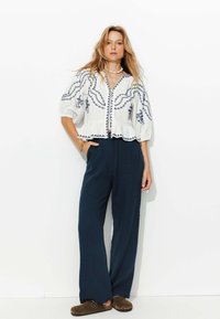 Blouse blanche brodée avec des motifs bleus, manches bouffantes et ourlet en peplum, associée à un pantalon large bleu marine et des chaussures brunes à enfiler.