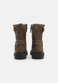 Bottines en daim marron avec un bout arrondi, des coutures contrastées, une fermeture éclair et des détails de sangle ajustable. Semelle en caoutchouc noire épaisse.