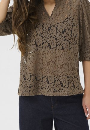 Žena v hnědém květovaném krajkovém topu s krátkými rukávy a tmavých džínových kalhotách, ruka opřená u obličeje, viditelné dlouhé vlnité vlasy.