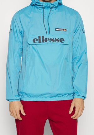 Man in turquoise ellesse windbreaker met capuchon en rode broek, met ontspannen handen langs de zij, zichtbaar van nek tot halverwege de dijen.