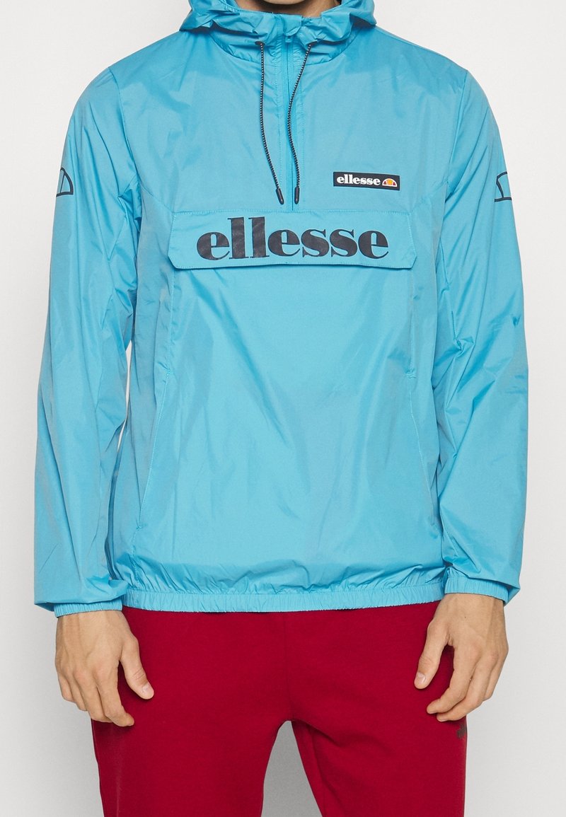 Hombre con chaqueta de chubasquero turquesa de ellesse con capucha y pantalones rojos, con las manos relajadas a los lados, visible desde el cuello hasta la mitad del muslo.
