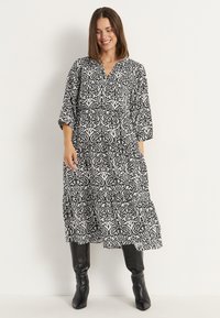 Schwarz-weiß gemustertes Kleid mit V-Ausschnitt, dreiviertel Ärmel und gestuftem Rock, ergänzt durch schwarze kniehohe Stiefel.