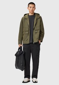 Veste à capuche vert olive avec deux poches avant, haut gris, pantalon cargo noir et baskets blanches. L'individu tient un sac noir.