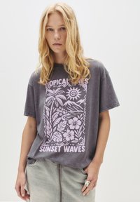 Grå oversized t-shirt med en pink tropisk grafik, der viser palmer, sol og teksten "SUNSET WAVES", parret med en grå denimskørt.