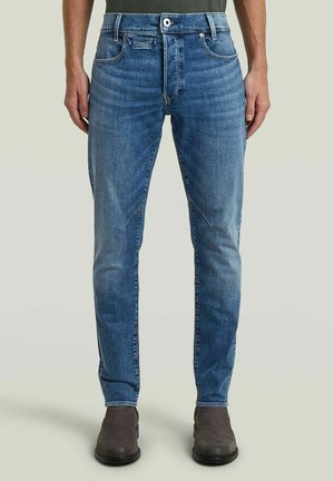 Homme portant un jean slim bleu avec une décoloration subtile et des bottines marron, debout devant un fond uni.