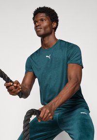 Teal trenings t-skjorte med rund hals og hvit Puma-logo, sammen med matchende slim-fit bukser, holder et svart treningsbånd.