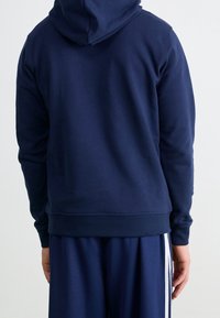 Marinblå hoodiesweatshirt tillverkad av bomullsblandat tyg. Har en avslappnad passform, ribbade muddkanter och en kenguruficka. Slät textur med minimal detaljering.