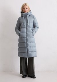 Lange lichtblauwe pufferjas met een hoge kraag, ritssluiting en twee zakken. Het materiaal lijkt glanzend met een horizontaal gequilt patroon.