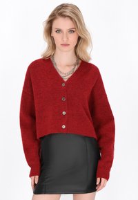 Rote, kurze Strickjacke mit V-Ausschnitt, langen Ärmeln und fünf Knöpfen. Hergestellt aus strukturiertem Strickmaterial. Kombiniert mit einem schwarzen Lederrock.