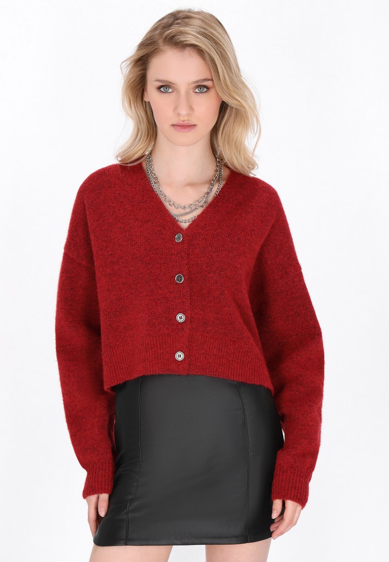 Rote, kurze Strickjacke mit V-Ausschnitt, langen Ärmeln und fünf Knöpfen. Hergestellt aus strukturiertem Strickmaterial. Kombiniert mit einem schwarzen Lederrock.