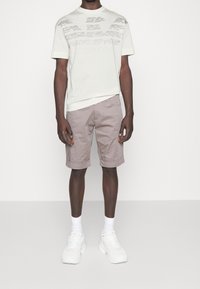 Uomo con una t-shirt grafica color avorio, pantaloni corti fino al ginocchio di un tenue colore malva, calze bianche e sneakers chunky bianche che si trova in piedi contro uno sfondo semplice.