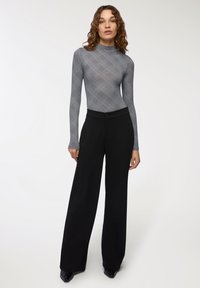 Donna con un top lungo a maniche grigio e modello, e pantaloni neri a gamba larga, in piedi con un'espressione neutra su uno sfondo bianco.