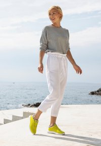 Grauer gerippter Pullover kombiniert mit weißen, zulaufenden Hosen und knallgelben Sneakers mit grauen Akzenten, stehend in der Nähe von Wasser an einem sonnigen Tag.