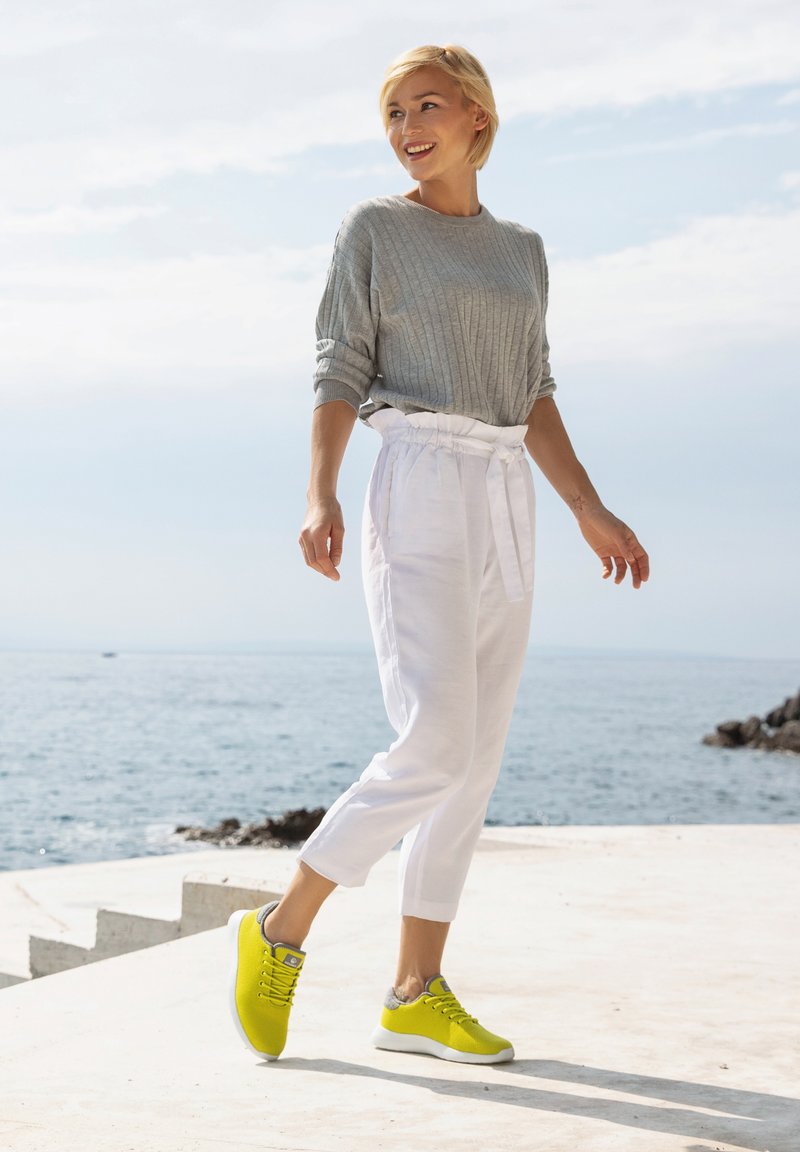 Grauer gerippter Pullover kombiniert mit weißen, zulaufenden Hosen und knallgelben Sneakers mit grauen Akzenten, stehend in der Nähe von Wasser an einem sonnigen Tag.