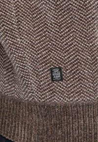 Tissu en tricot chevron marron et beige avec une petite étiquette rectangulaire noire "Ritchie Jeans" cousue près de l'ourlet côtelé.