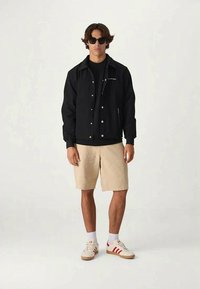 Young man wearing black jacket, beige shorts, white sneakers with red stripes, white socks, and black sunglasses, standing against plain background.