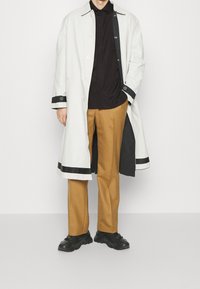 Trench coat lungo di un grigio chiaro con accenti neri, camicia nera e pantaloni beige. Sneakers nere completano l'outfit. Design minimalista.