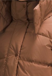 Chaqueta acolchada marrón con una textura suave y lisa. Presenta grandes secciones acolchadas y botones a presión marrones para el cierre.