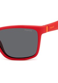 Occhiali da sole in plastica rossa con finitura opaca, lenti grigie scure e marchio Polaroid sulla parte superiore sinistra delle lenti e sull'asta. Accento arancione sulla cerniera.