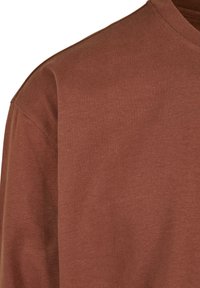 Camiseta de algodón marrón con cuello redondo y textura acanalada. Costuras visibles a lo largo de las uniones. La tela parece suave y casual.