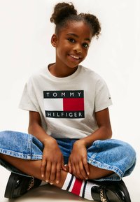 Szara koszulka z czarną, czerwoną i białą grafiką z napisem "TOMMY HILFIGER". Połączona z niebieskimi dżinsami w szerokim kroju i czarnymi butami z łańcuchem.