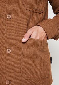 Brown textured jacket with buttons. Features a patch pocket, ribbed fabric, and a tag with "EST 1993" on the side.