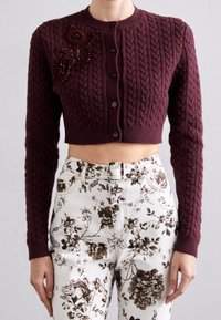 Bordeauxfarbene, kurz geschnittene Strickjacke mit geripptem Muster, verziert mit einer mit Perlen besetzten Blumenakzent auf der linken Schulter und sechs Knöpfen vorne. Darunter floral gemusterte Hosen.