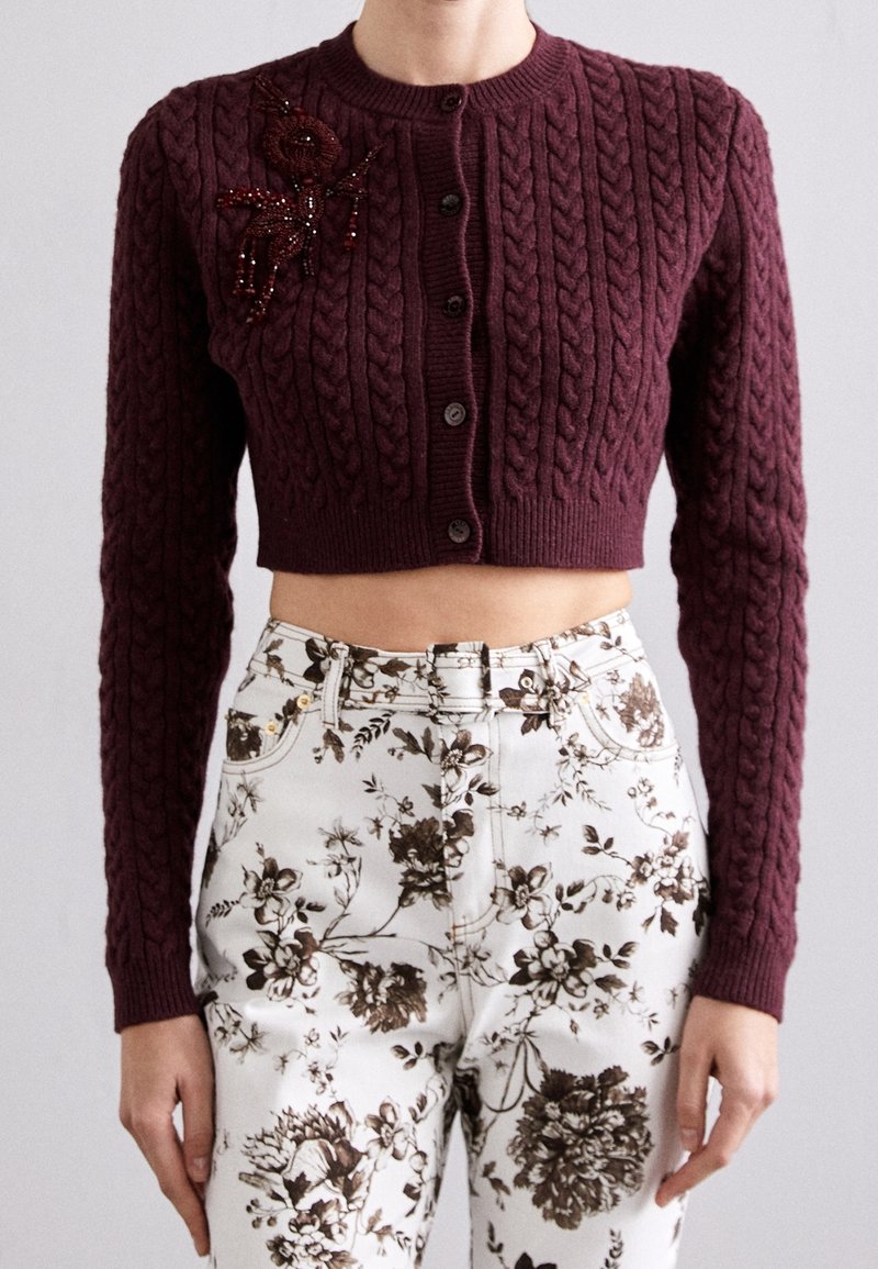 Bordeauxfarbene, kurz geschnittene Strickjacke mit geripptem Muster, verziert mit einer mit Perlen besetzten Blumenakzent auf der linken Schulter und sechs Knöpfen vorne. Darunter floral gemusterte Hosen.