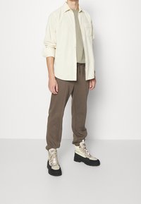 Camicia button-up color crema chiaro sopra una maglietta color taupe, abbinata a pantaloni jogger marroni e stivali da escursione beige con suole nere.