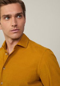 Camisa mostaza de botones con un cuello clásico, tejido texturizado y dos botones visibles en el placket. Modelo con fondo neutro.