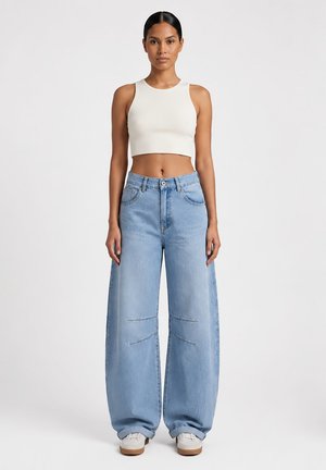 Kobieta stojąca przodem, ubrana w biały bezrękawnik typu crop top, szerokie jasnoniebieskie dżinsy i białe trampki na jednolitym tle.