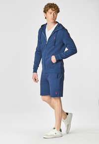 Felpa con zip color navy e tasche, abbinata a pantaloni corti coordinati. T-shirt bianca a scollo a V sotto. Il modello indossa scarpe da ginnastica bianche. Dettaglio logo rosso.